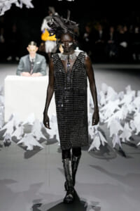 Runway model in a black beaded dress with a large sculptural black headpiece and metallic jewelry, walking a white-lit catwalk.