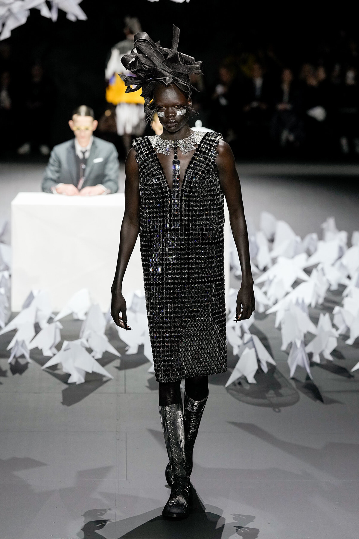Runway model in a black beaded dress with a large sculptural black headpiece and metallic jewelry, walking a white-lit catwalk.