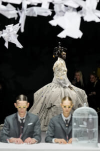 Model in a voluminous silver plaid ball gown walks the runway beneath hanging white paper birds.
