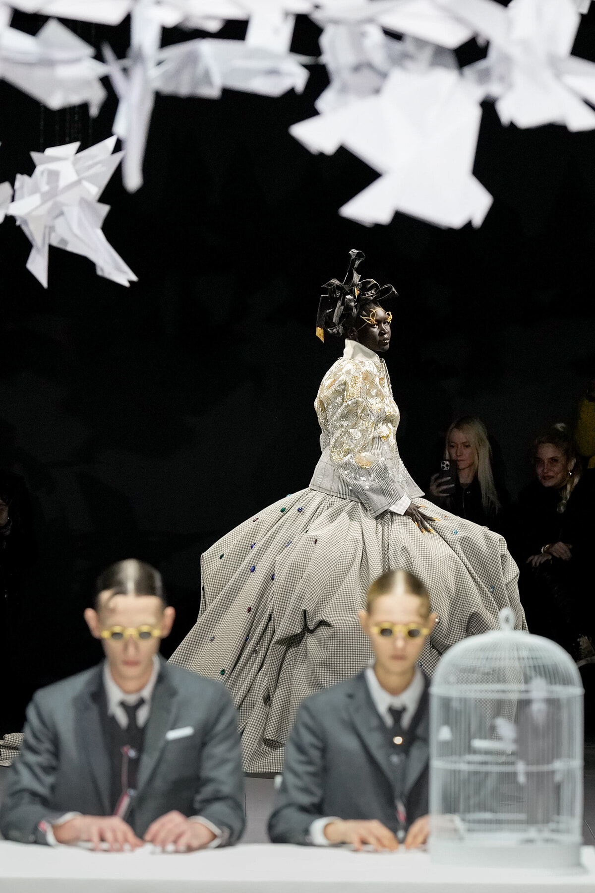 Model in a voluminous silver plaid ball gown walks the runway beneath hanging white paper birds.