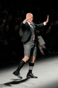 A formally dressed older man walks a runway in gray suit jacket and shorts, with striped socks, raising both hands in a gesture toward the audience.