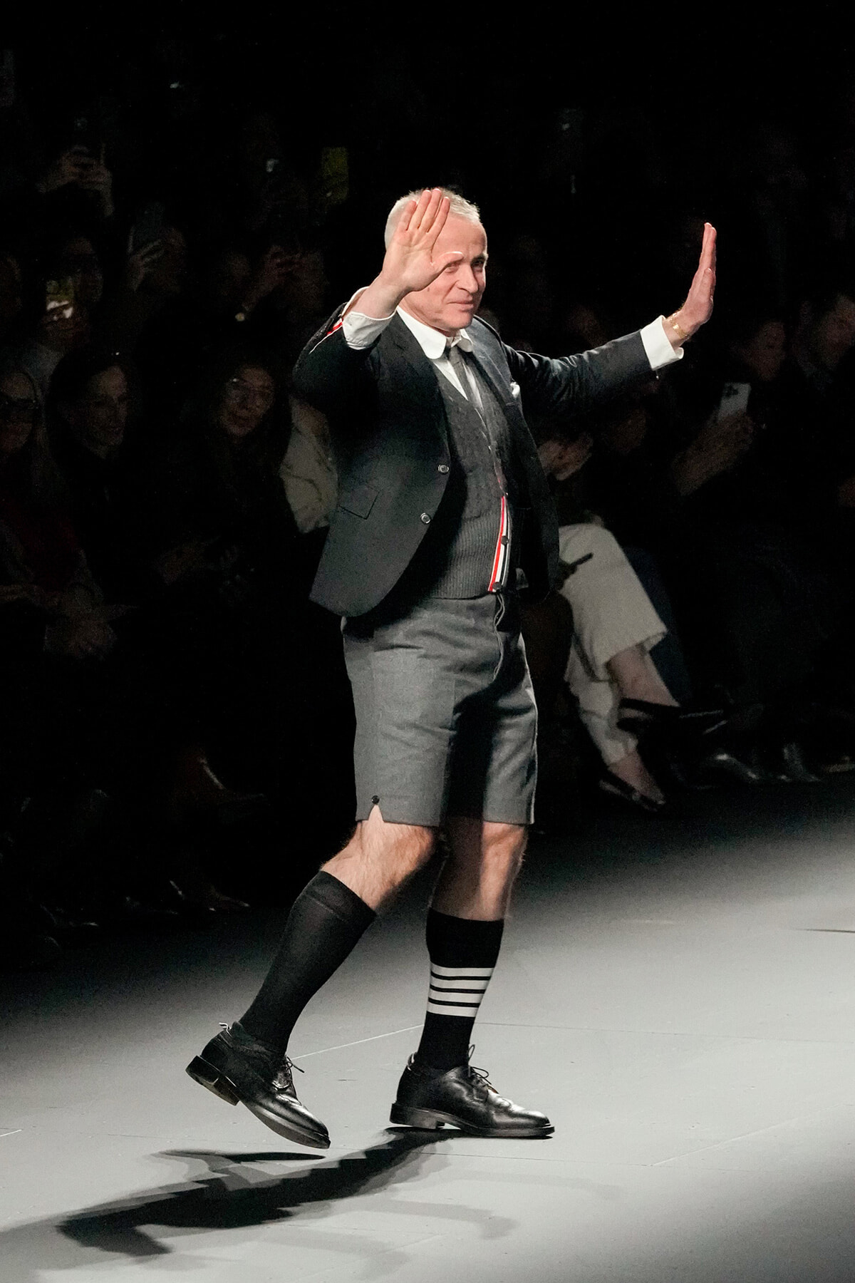 A formally dressed older man walks a runway in gray suit jacket and shorts, with striped socks, raising both hands in a gesture toward the audience.
