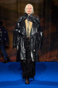 Model walks the runway in an oversized black leather trench over a floor-length beaded black gown on a blue catwalk