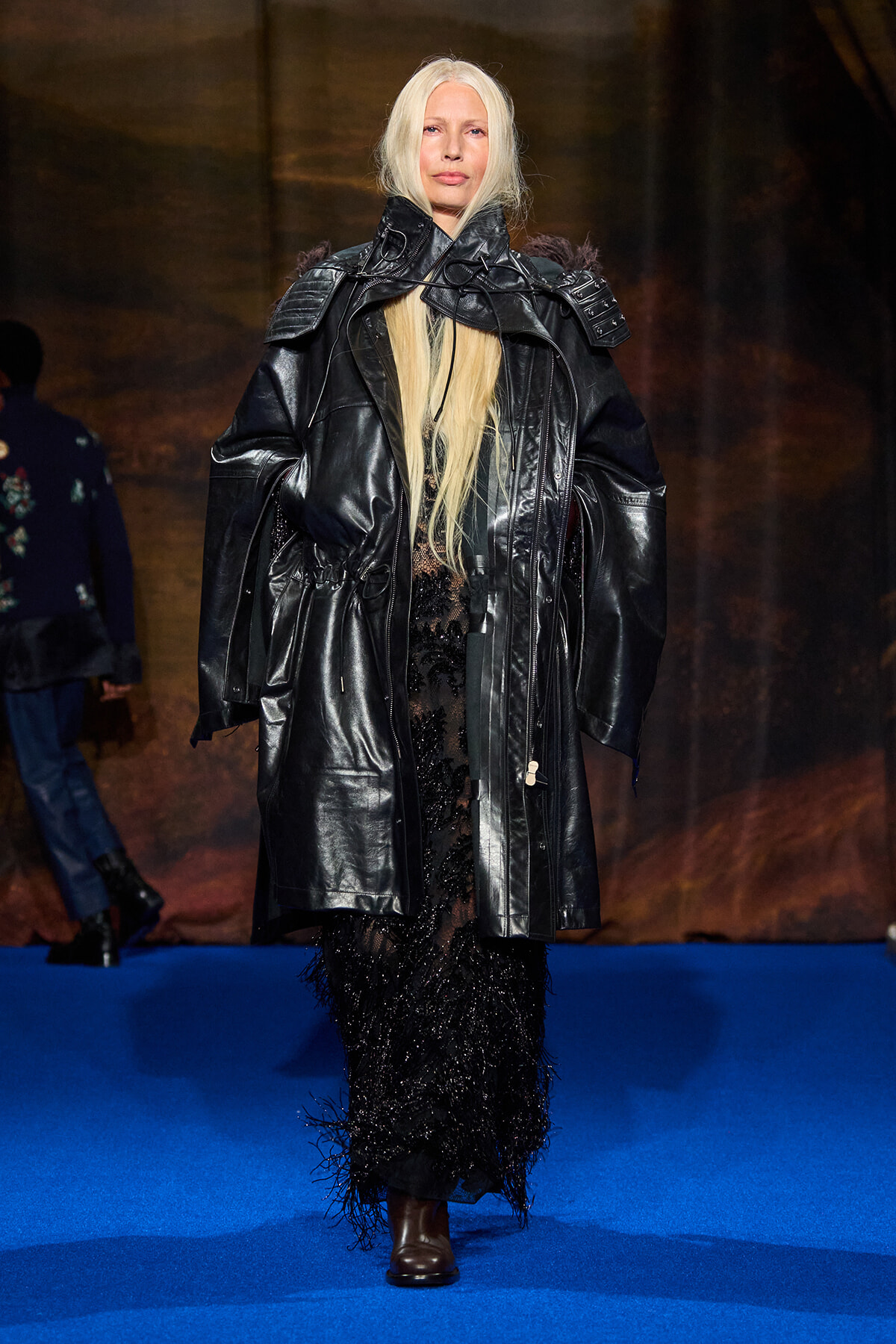Model walks the runway in an oversized black leather trench over a floor-length beaded black gown on a blue catwalk