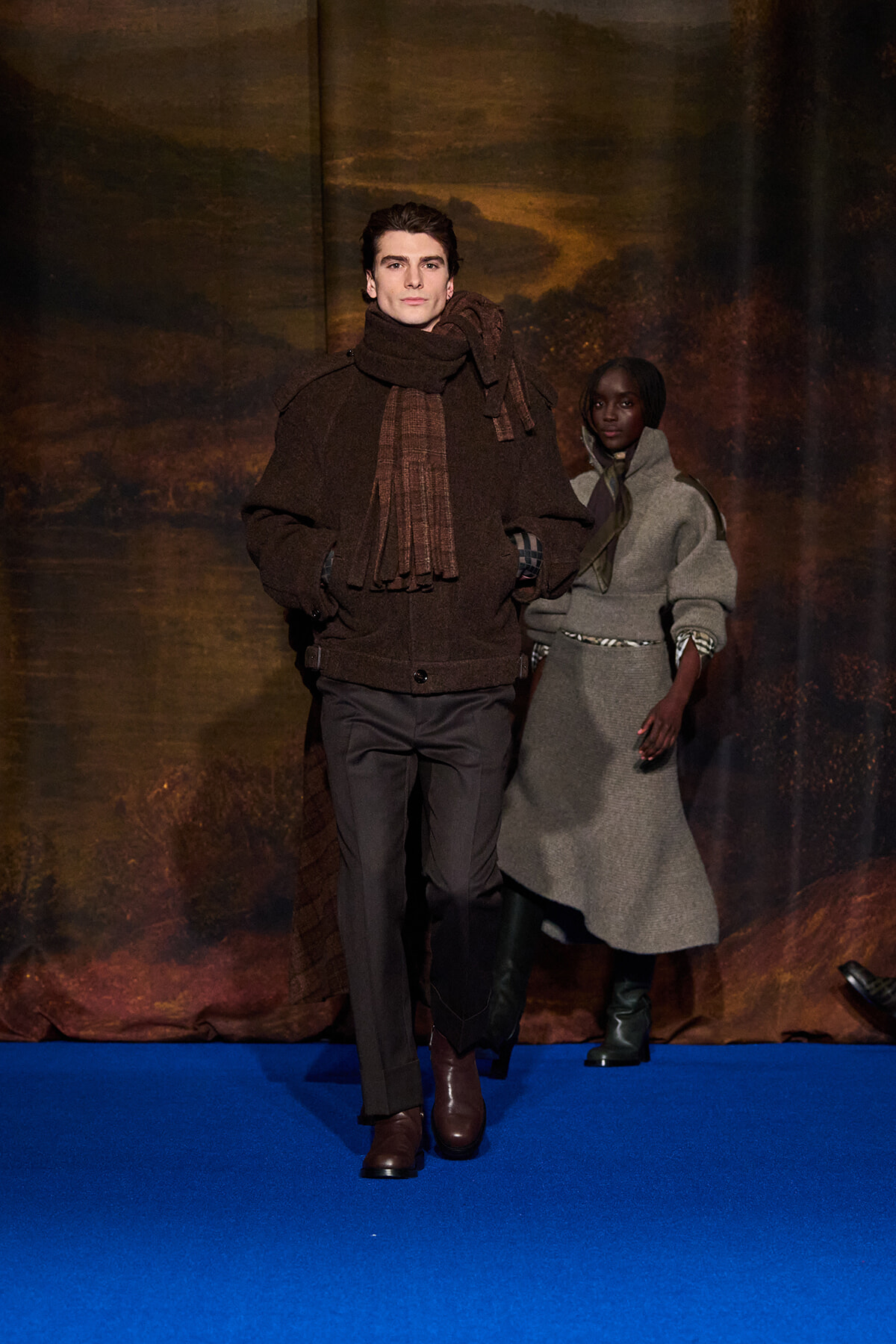 Male model in a brown coat and scarf walks the runway with hands in pockets, a female model in a gray coat follows behind on a blue catwalk.