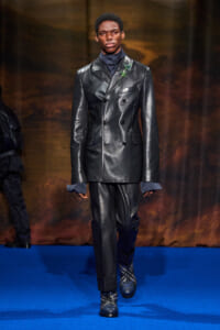 Male model on a blue runway wearing a glossy black double-breasted leather suit with a green boutonniere.