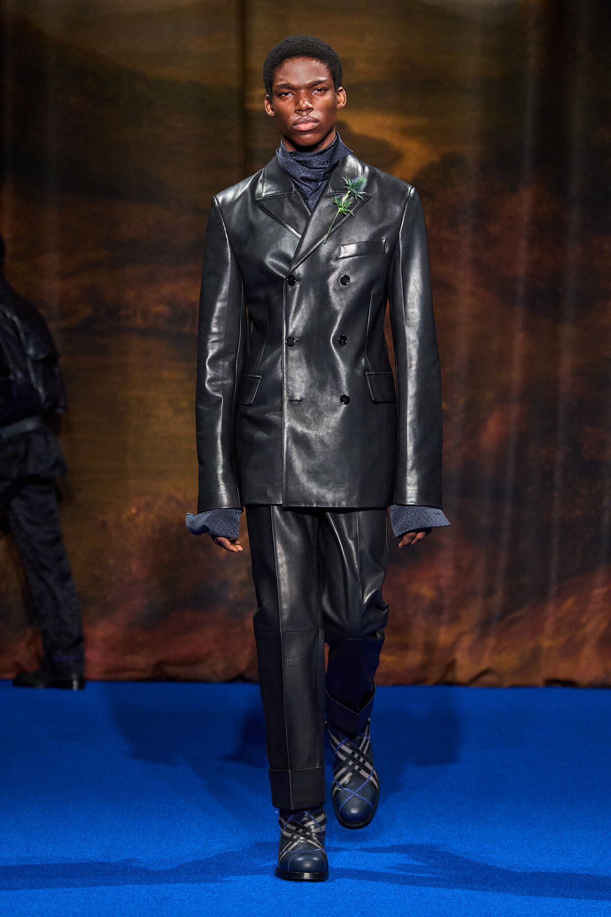 Male model on a blue runway wearing a glossy black double-breasted leather suit with a green boutonniere.