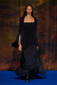 Model walks a runway in a long black ruched gown with fringe sleeves and layered fringe skirt, against a dark backdrop.