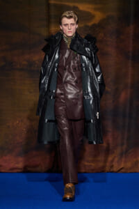 Male model on a blue runway wearing a burgundy double‑breasted leather suit under a long black coat; brown lace‑up boots visible .