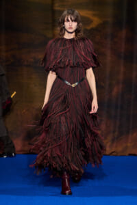 Model walking a burgundy fringe gown with layered, feather-like strips on a blue runway.