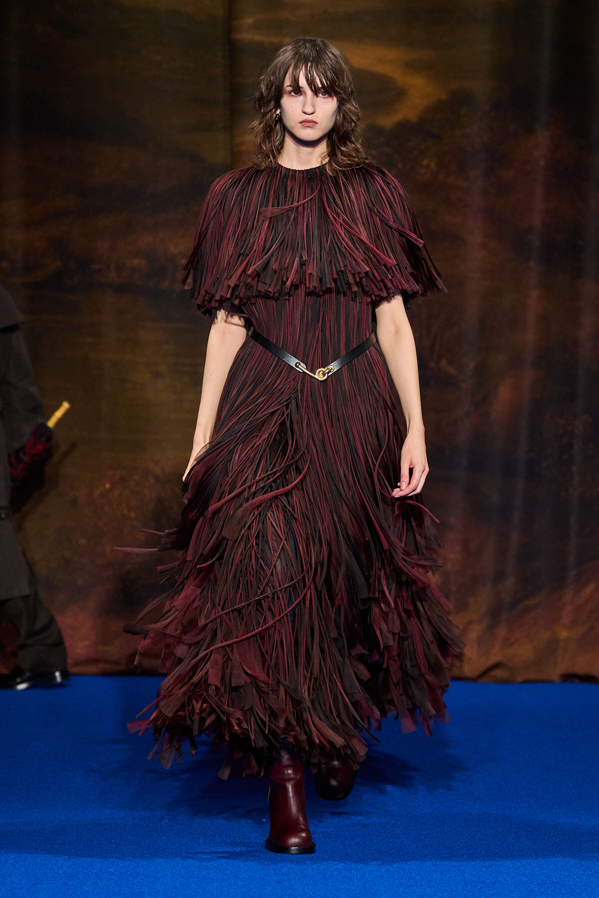Model walking a burgundy fringe gown with layered, feather-like strips on a blue runway.