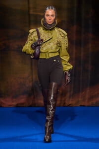 Runway model walking in an olive-green embroidered jacket, black pants, tall brown boots, gloves, and a headscarf.