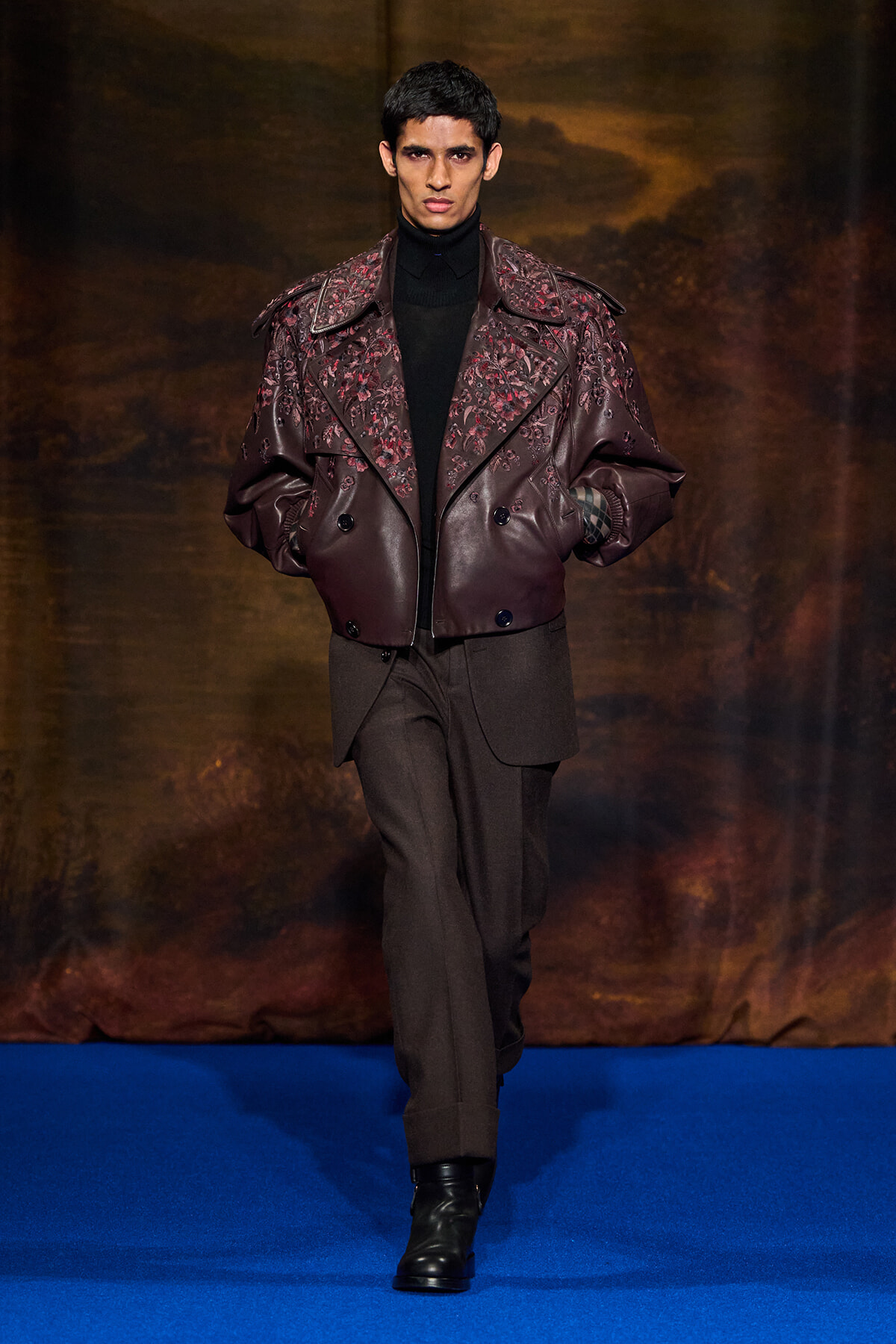 Male model walks a runway wearing a burgundy embroidered leather jacket over a black turtleneck, brown trousers, and black boots on a blue catwalk backdrop.