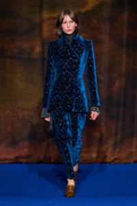 Model walking a blue velvet patterned suit on a runway, wearing a dark turtleneck and brown loafers.