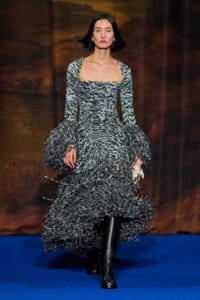 Model walking a runway in a gray textured gown with feathered sleeves and ruffled hem, black knee-high boots.