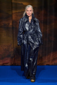 Older woman on a blue runway wearing a shiny navy trench coat with belt and ruffled collar, looking at camera.