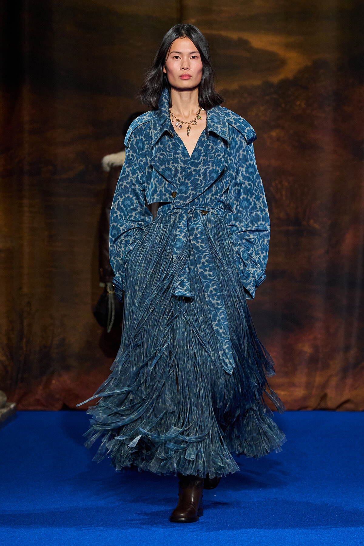 Model walks a blue-patterned trench with a matching fringed skirt on a blue runway, wearing brown ankle boots and a gold necklace