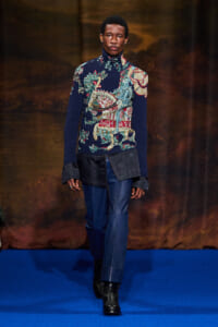 Male model walking a runway in a dark blue knit turtleneck with a colorful animal/landscape pattern, paired with blue pants and black boots.