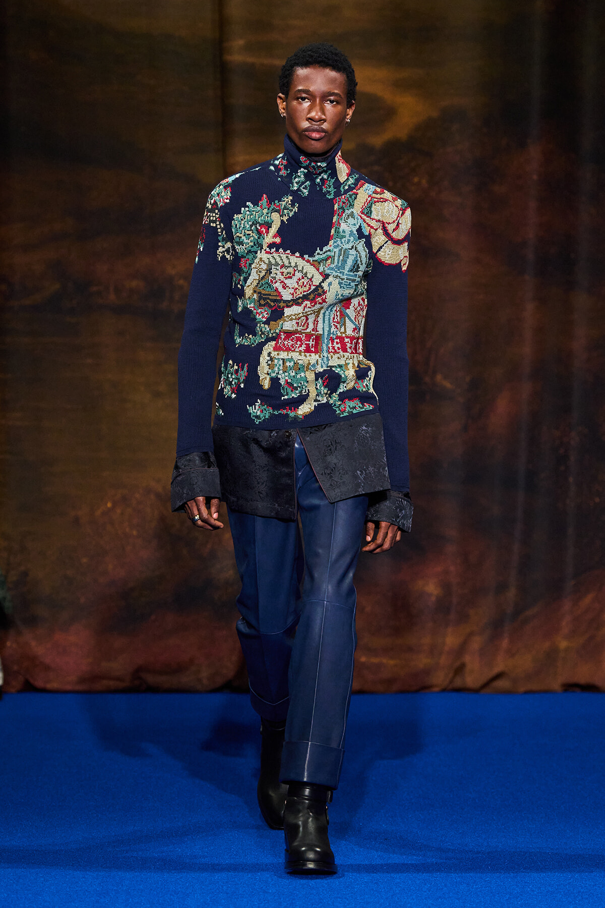 Male model walking a runway in a dark blue knit turtleneck with a colorful animal/landscape pattern, paired with blue pants and black boots.