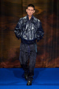 Male model walks the runway in a dark glossy trench coat with blue piping and belt.