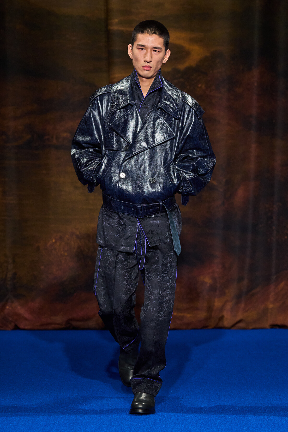 Male model walks the runway in a dark glossy trench coat with blue piping and belt.