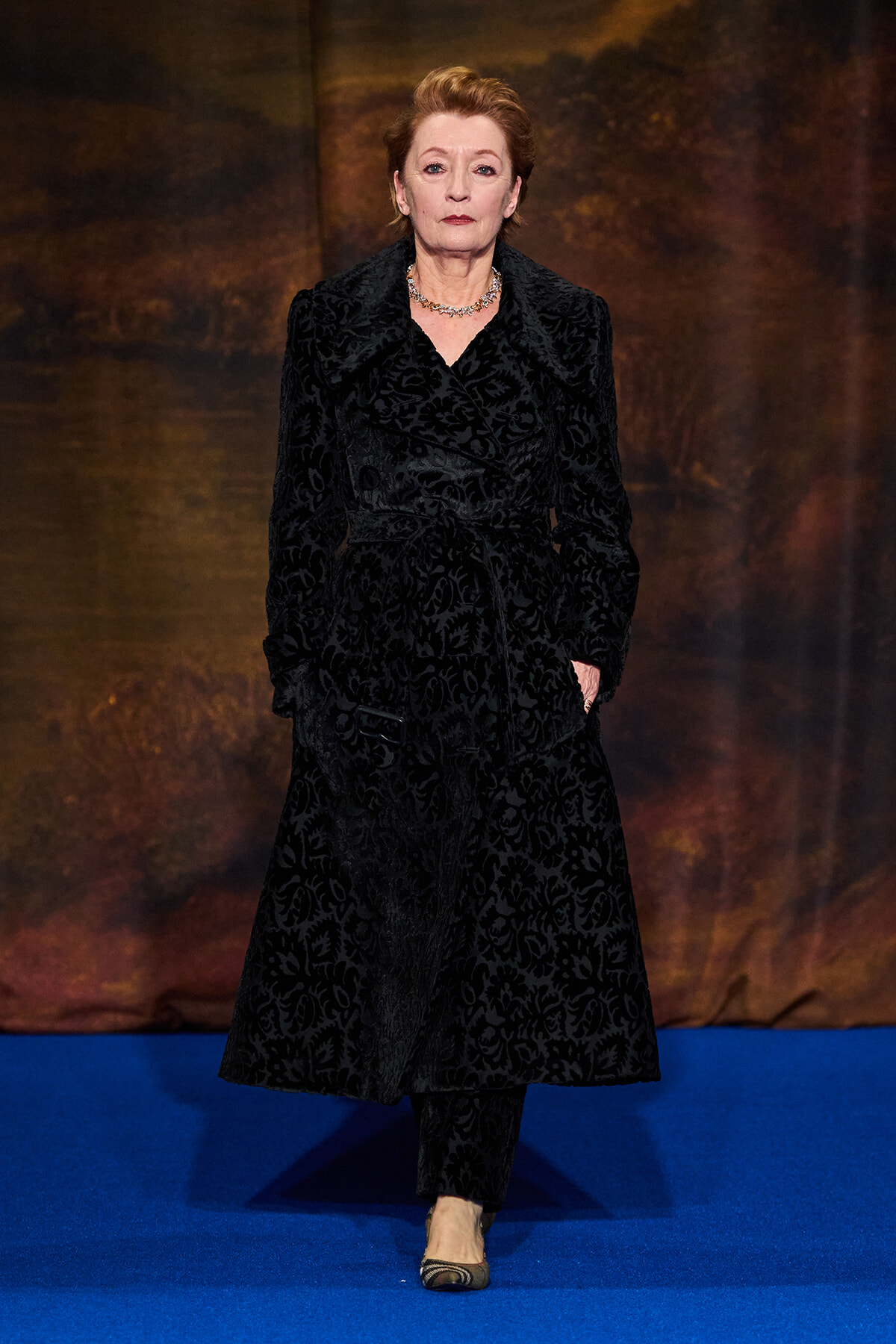 Older woman wearing a long black patterned velvet coat, matching pants, and a silver necklace walks down a blue carpet runway.