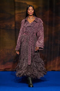 Model wearing a purple animal-print trench with a fringed, layered skirt walking down a blue runway.