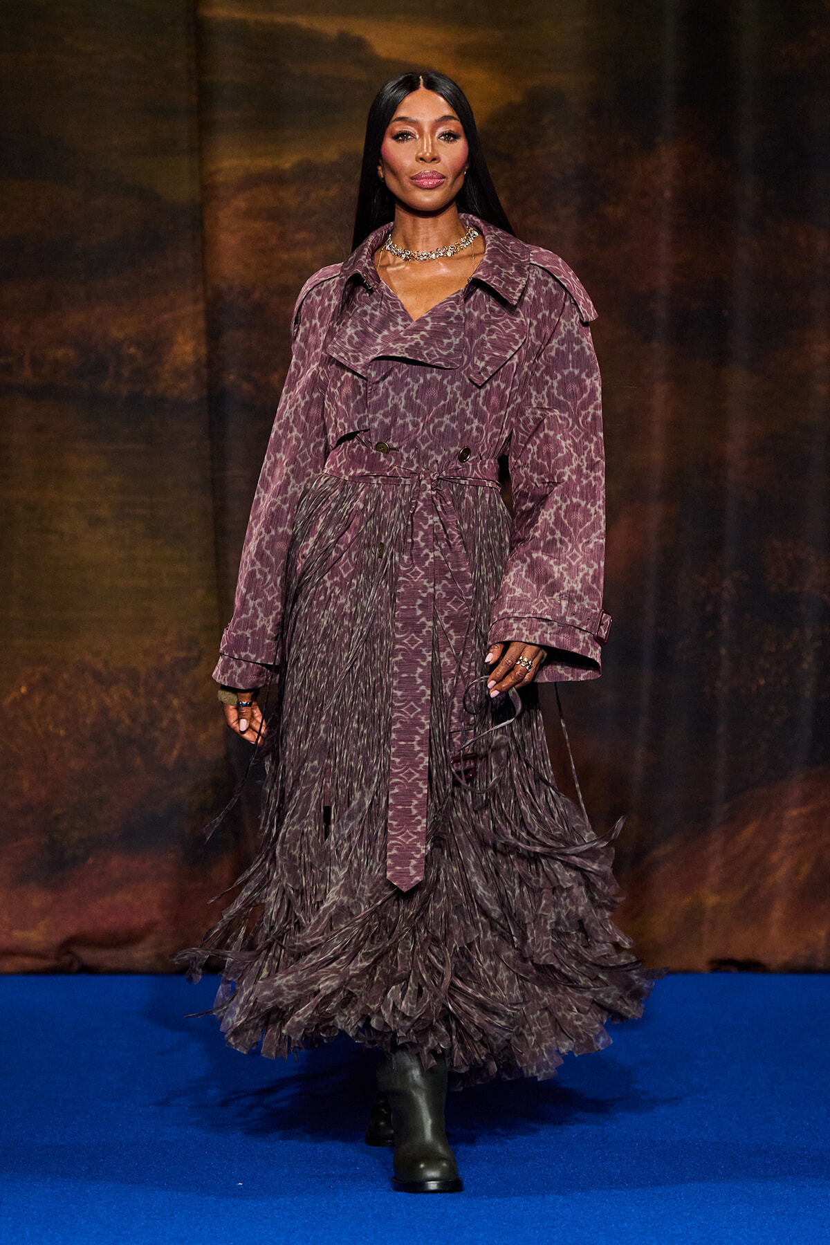 Model wearing a purple animal-print trench with a fringed, layered skirt walking down a blue runway.