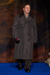 Man in a long gray trench coat, scarf, and gloves stands on a blue carpet, holding a red item in his right hand on a stage backdrop.