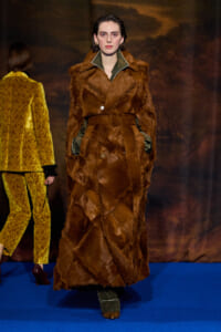 Model on a blue runway wearing a long brown fur coat with a green-trimmed collar and gloves.