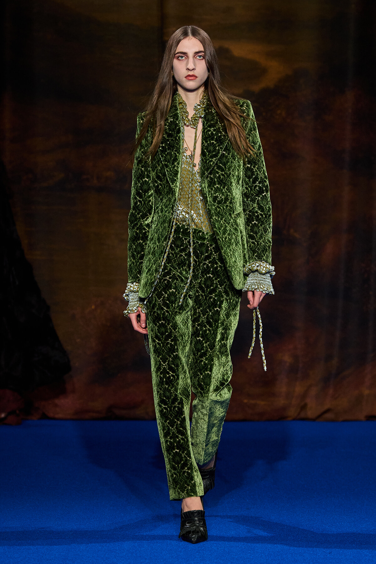 Model walking the runway in a green velvet suit with a patterned blazer and matching trousers, on a blue catwalk.