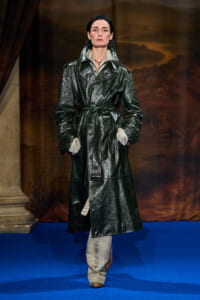 Model on a fashion runway wearing a glossy dark green leather trench coat with a belt, hands in pockets on a blue carpeted catwalk.