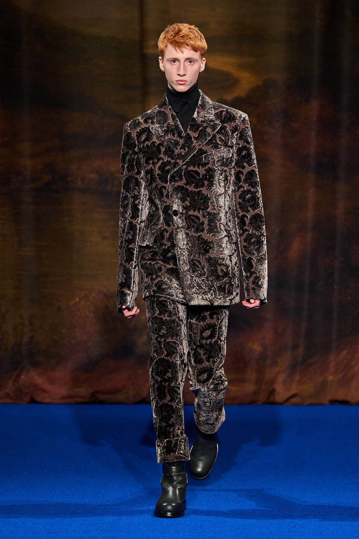 Male model wearing a dark velvet floral suit with a black turtleneck, walking a blue runway.