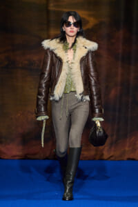 Model on a runway wearing a brown shearling jacket with cream fur trim, green blouse, gray knit pants, black knee-high boots, sunglasses, and a black handbag.
