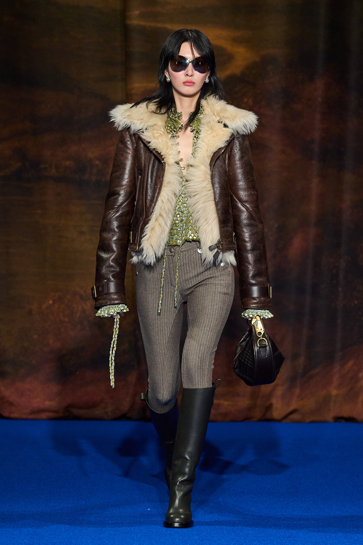 Model on a runway wearing a brown shearling jacket with cream fur trim, green blouse, gray knit pants, black knee-high boots, sunglasses, and a black handbag.