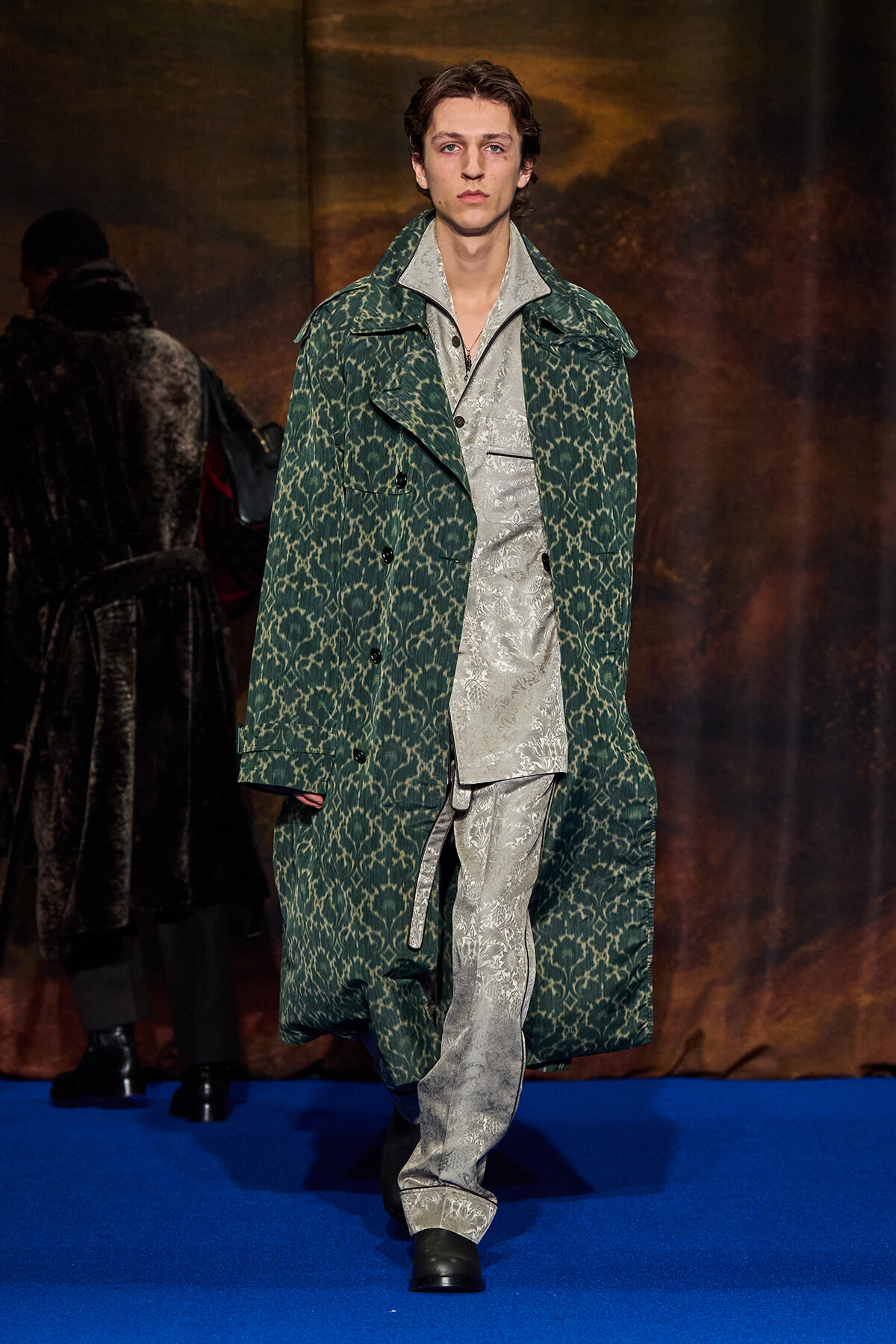 Male model on a runway wearing a green patterned long coat over a silver damask suit on a blue catwalk surface.
