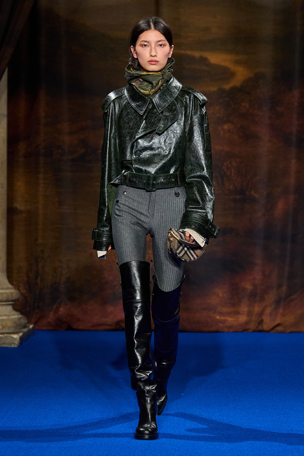 Model on a blue runway wearing a glossy dark leather trench, gray pants, tall black boots, and a plaid clutch purse.