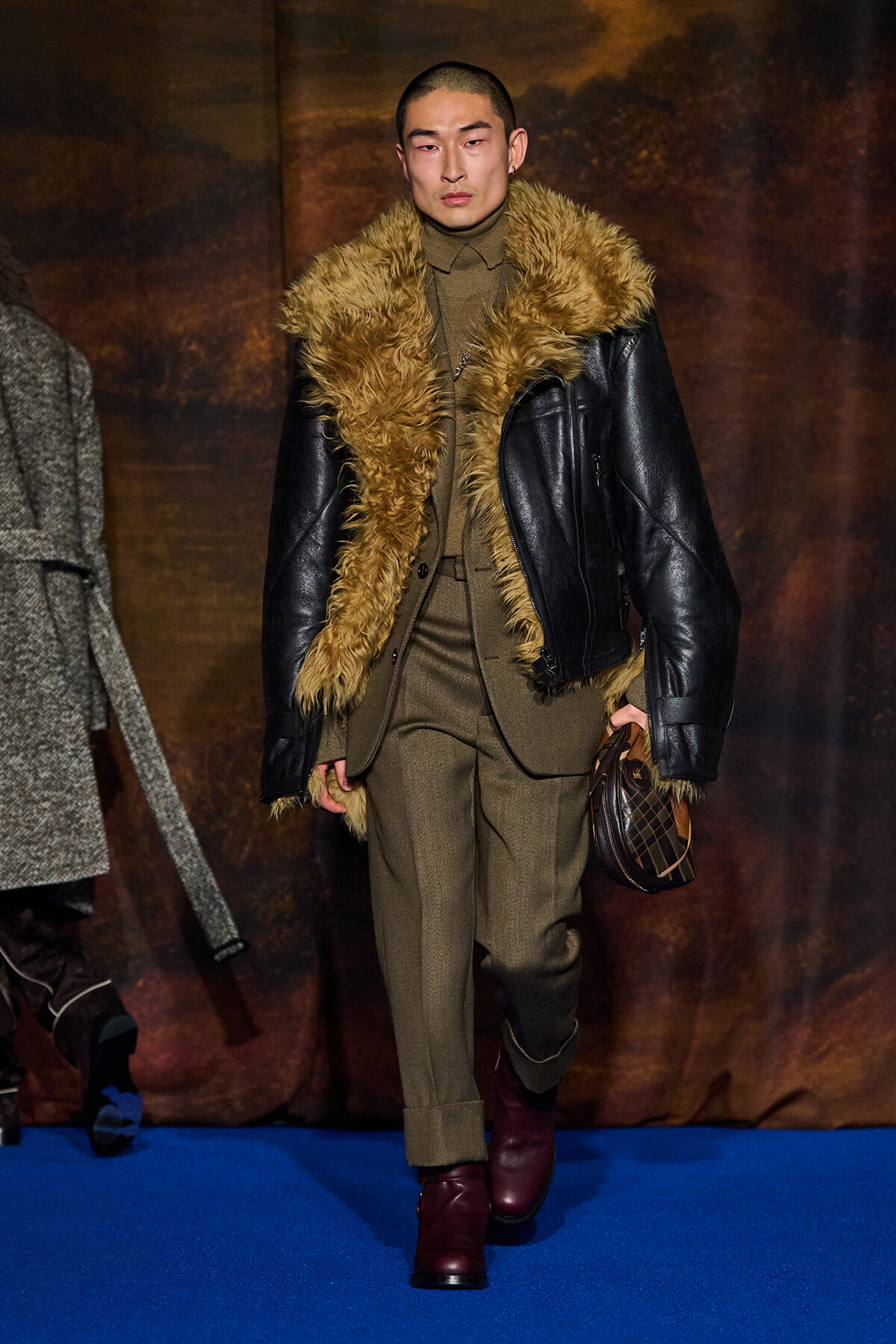 Male model wearing an olive-brown suit with a fur-trimmed black leather jacket, burgundy boots, and a small plaid bag on a blue runway.