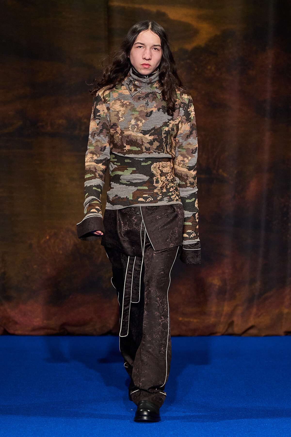 Model walking a fashion runway in a camouflage-knit turtleneck sweater and dark patterned pants with piping.