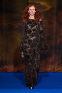 Model with curly red hair walks a runway in a long black lace gown with beadwork and fringe against a dark backdrop on a blue catwalk edge.