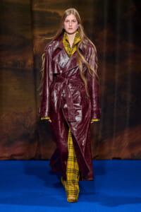 Model walking the runway in a burgundy leather trench with a matching belt over a yellow plaid outfit, against a blue catwalk backdrop.