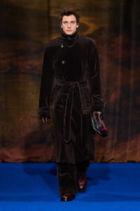 Male model walking a runway in a dark velvet trench coat with belt, burgundy gloves, and ankle boots, holding a plaid cap in one hand.