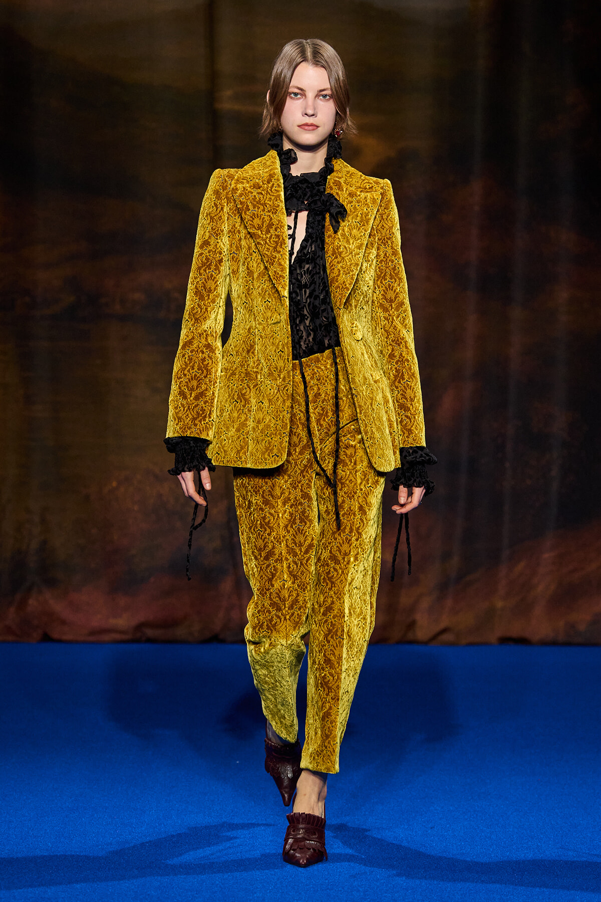 Model walks the runway in a gold-yellow velvet suit with a black lace blouse and bow accents, on a blue catwalk surface.