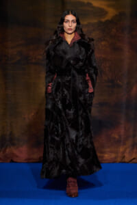 Model on a blue runway wearing a long black fur coat with burgundy lining and cuffs.