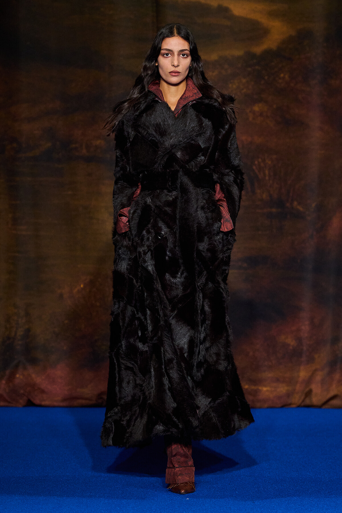 Model on a blue runway wearing a long black fur coat with burgundy lining and cuffs.