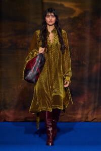 Model walking on a blue runway wearing a long yellow patterned dress with burgundy knee-high boots and a large maroon and black shoulder bag.