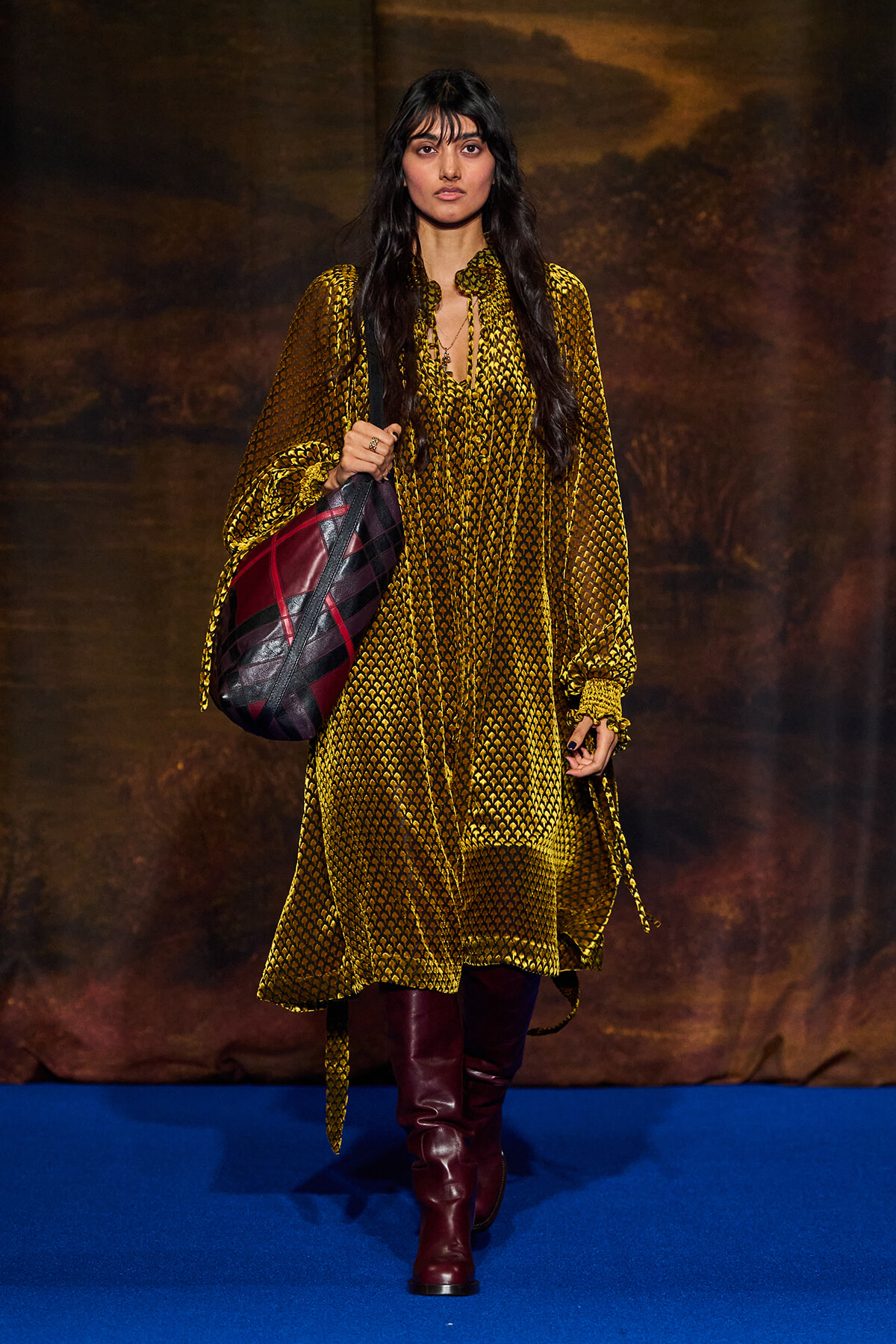 Model walking on a blue runway wearing a long yellow patterned dress with burgundy knee-high boots and a large maroon and black shoulder bag.