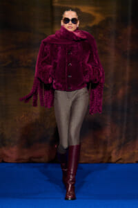 Model on a blue runway wearing a oversized burgundy faux-fur coat, gray ribbed pants, burgundy knee-high boots, and dark sunglasses.