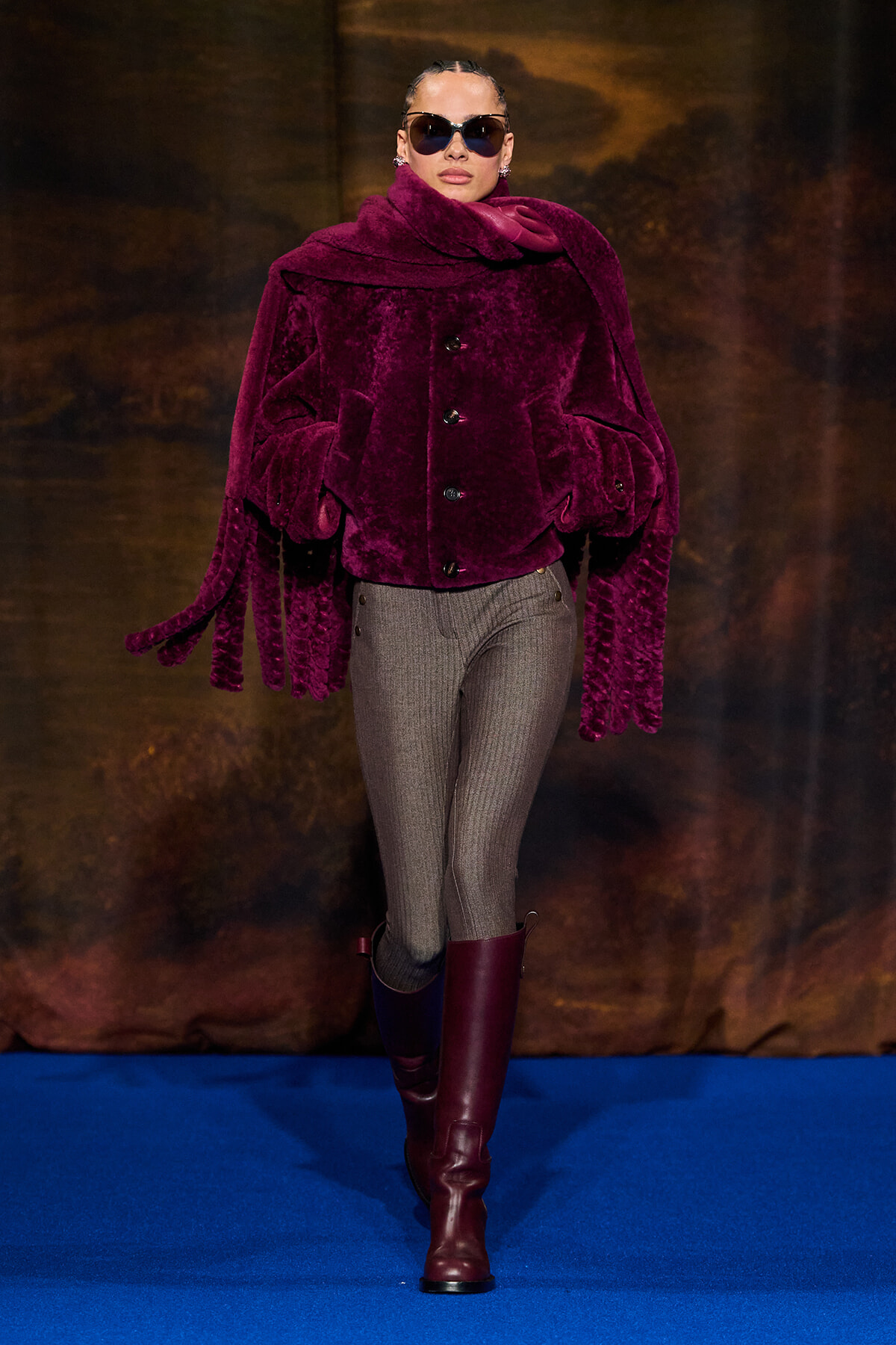 Model on a blue runway wearing a oversized burgundy faux-fur coat, gray ribbed pants, burgundy knee-high boots, and dark sunglasses.