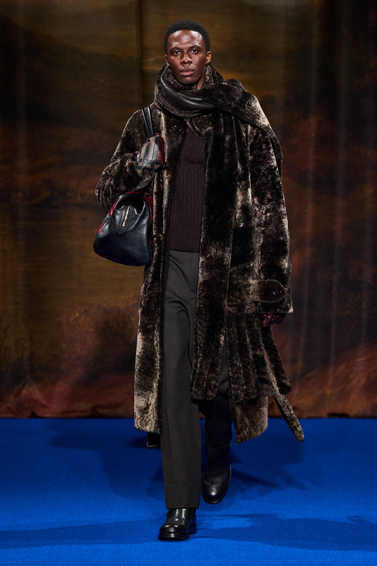 Model walks a fashion show wearing a long dark fur coat, black sweater, and trousers, carrying a black leather bag with plaid accents on a blue runway.